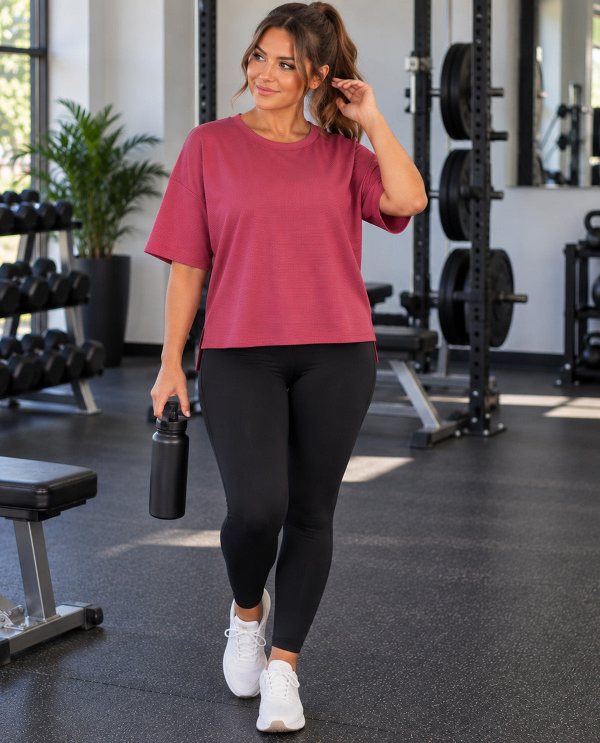A woman in a gym wearing a dusty rose short-sleeve t-shirt, black leggings, and white sneakers, holding a black water bottle.