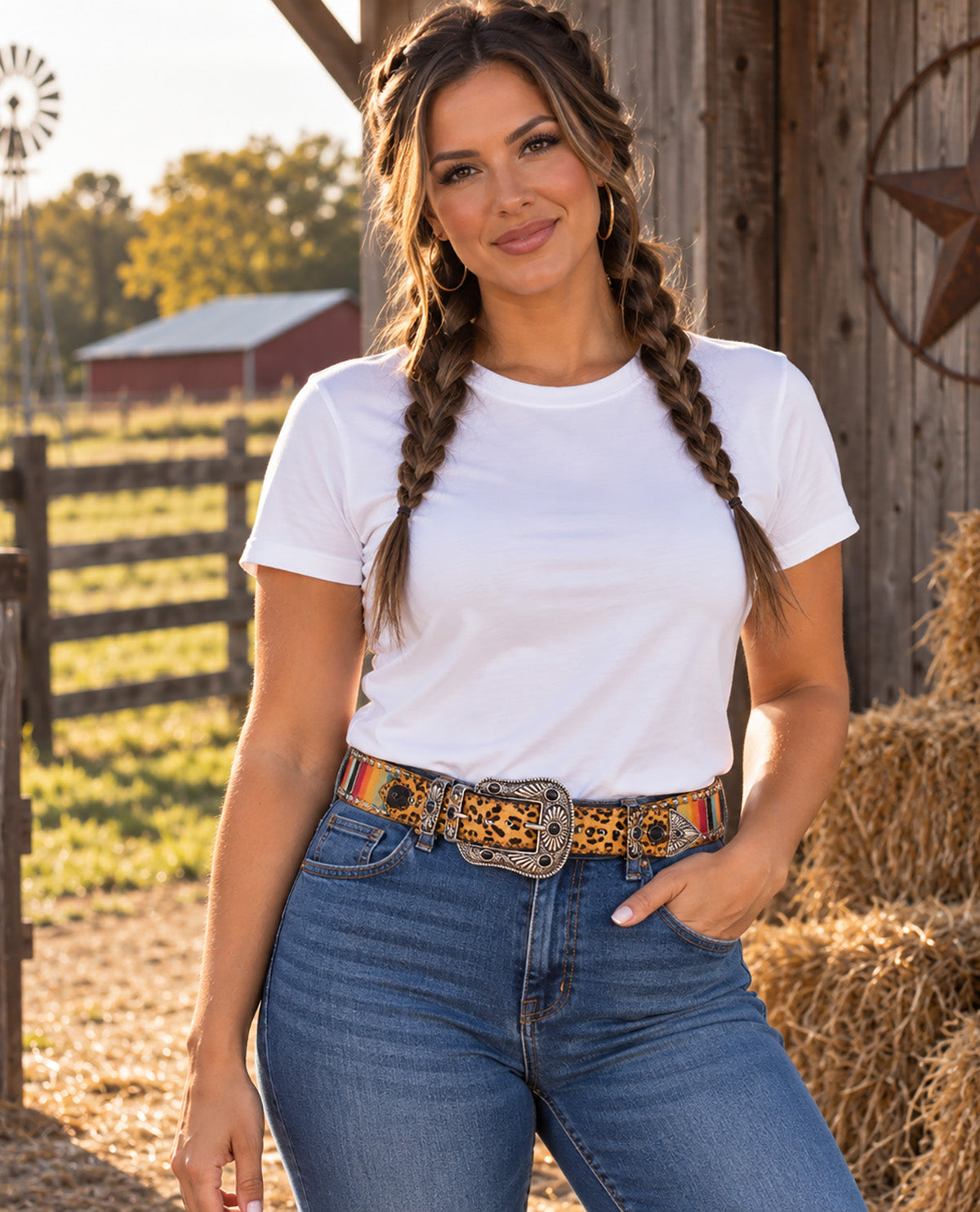 A woman posing outdoors in a white crewneck t-shirt and blue jeans, accessorized with a leopard print belt featuring colorful western-style trim and a large silver buckle. The background shows a rustic barn, hay bale, and fence.