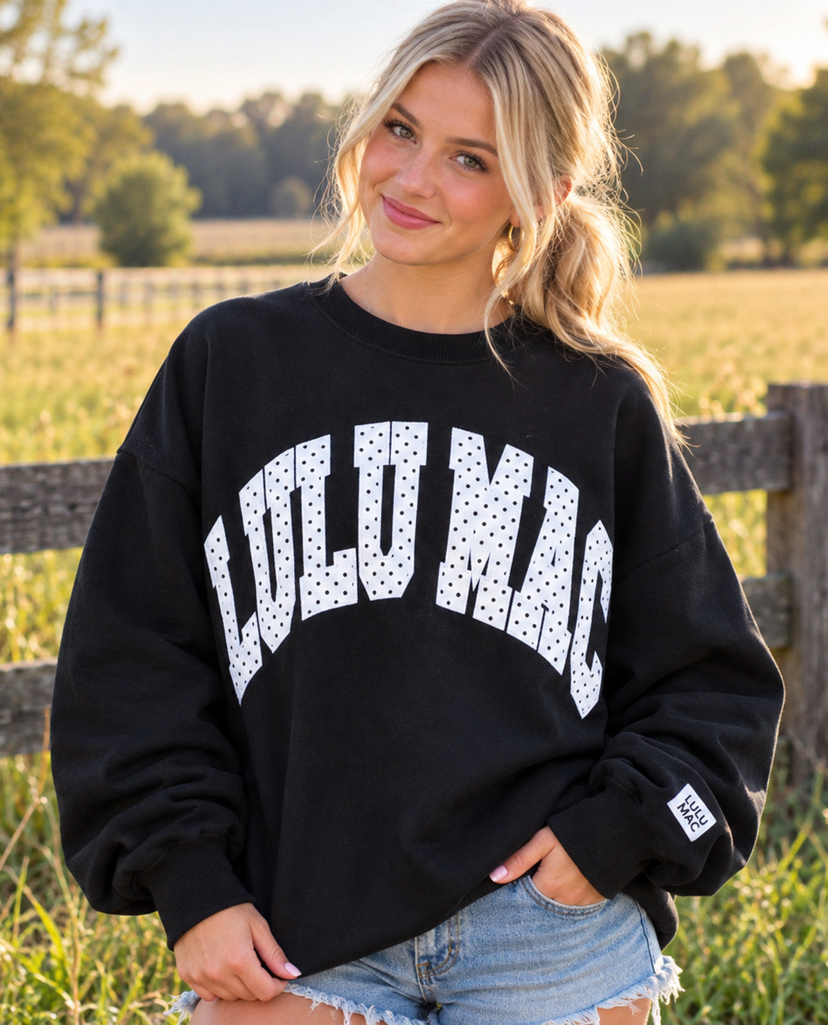 A woman wearing a black crewneck sweatshirt with 'LULUMAC' printed in white dotted letters across the front, paired with light blue denim shorts, set against a blurred outdoor background of a field and fence.