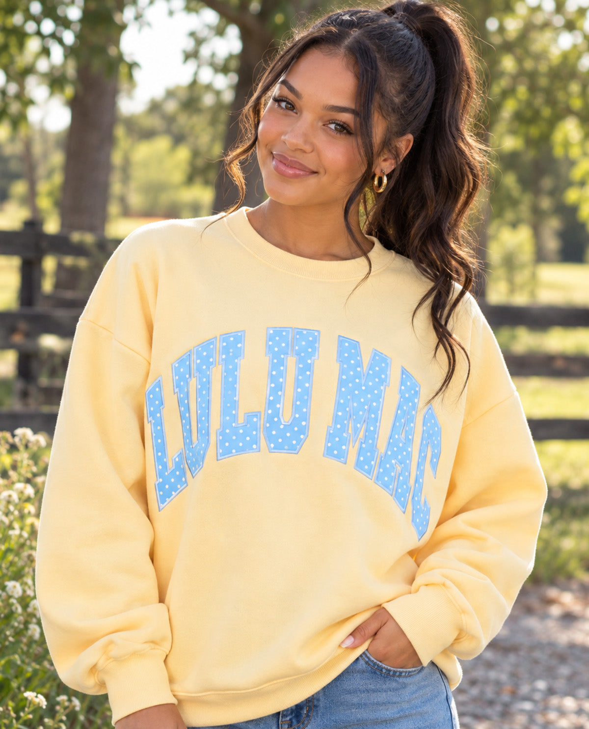 A woman wearing a pale yellow crewneck sweatshirt with 'LULU MAC' printed in blue dotted letters across the chest, standing outdoors in front of a blurred fence and trees.