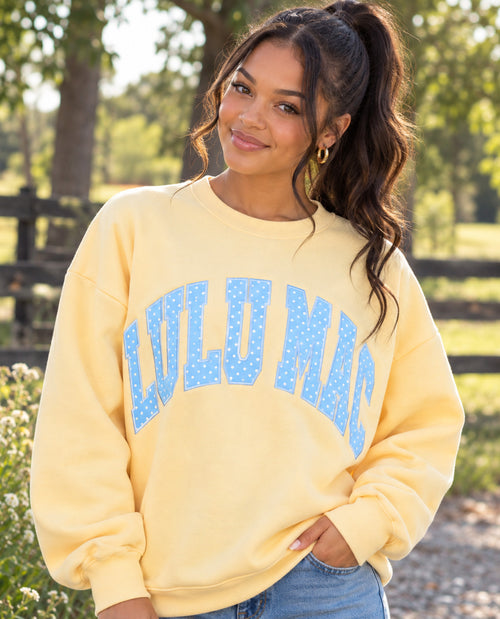 A woman wearing a pale yellow crewneck sweatshirt with 'LULU MAC' printed in blue dotted letters across the chest, standing outdoors in front of a blurred fence and trees.