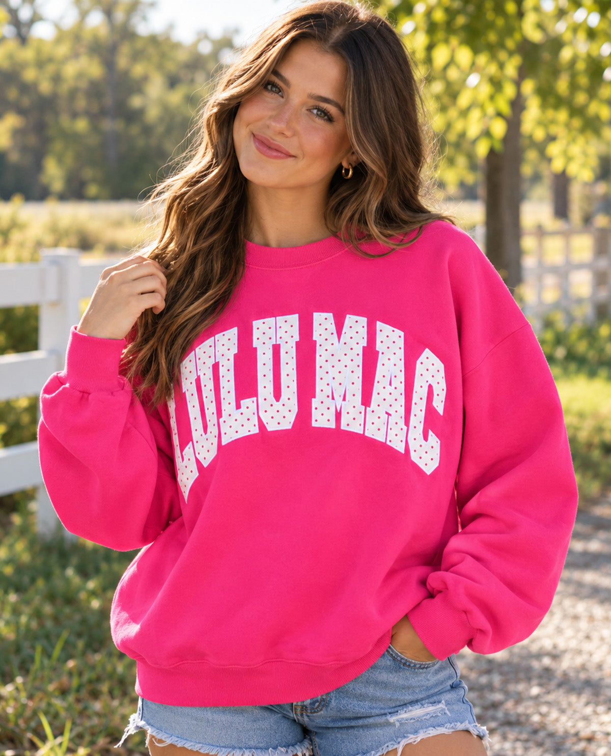 A woman wearing a bright pink crewneck sweatshirt with 'LULUMAC' printed in white arched letters with tiny polka dots, paired with denim shorts, set against a blurred outdoor background of trees and a white fence.