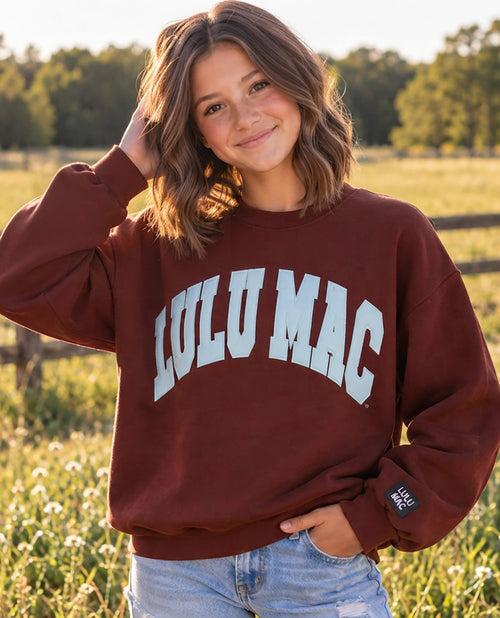 A woman wearing a maroon crewneck sweatshirt with 'LULU MAC' printed in white arched letters on the front, standing in a grassy field.