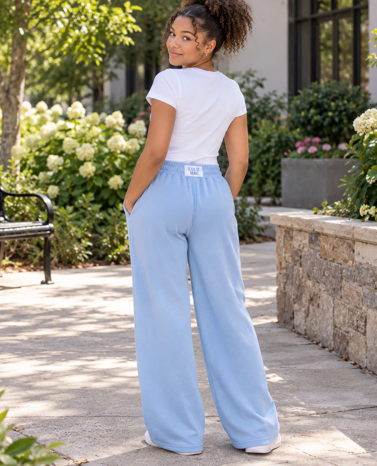 A woman wearing light blue wide-leg sweatpants and a white t-shirt, photographed from behind in an outdoor garden setting.