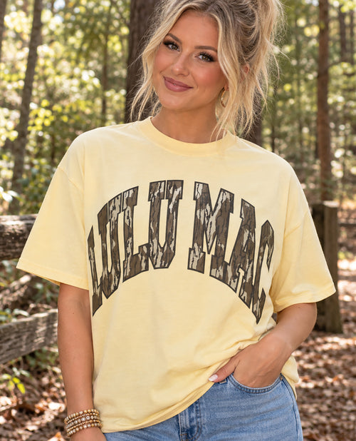 A woman wearing a pale yellow graphic t-shirt with 'LULU MAC' printed in distressed black letters, set against a blurred forest background.
