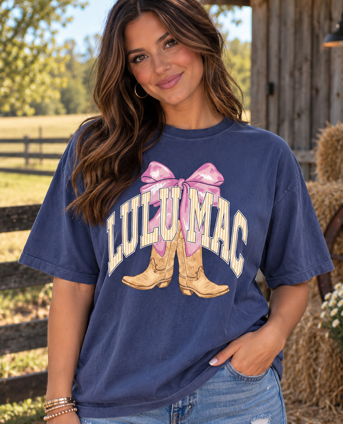 A woman wearing a navy blue graphic t-shirt with a pink bow and cowboy boots design that reads 'LULU MAC' in white arched letters, set against a rustic outdoor background.