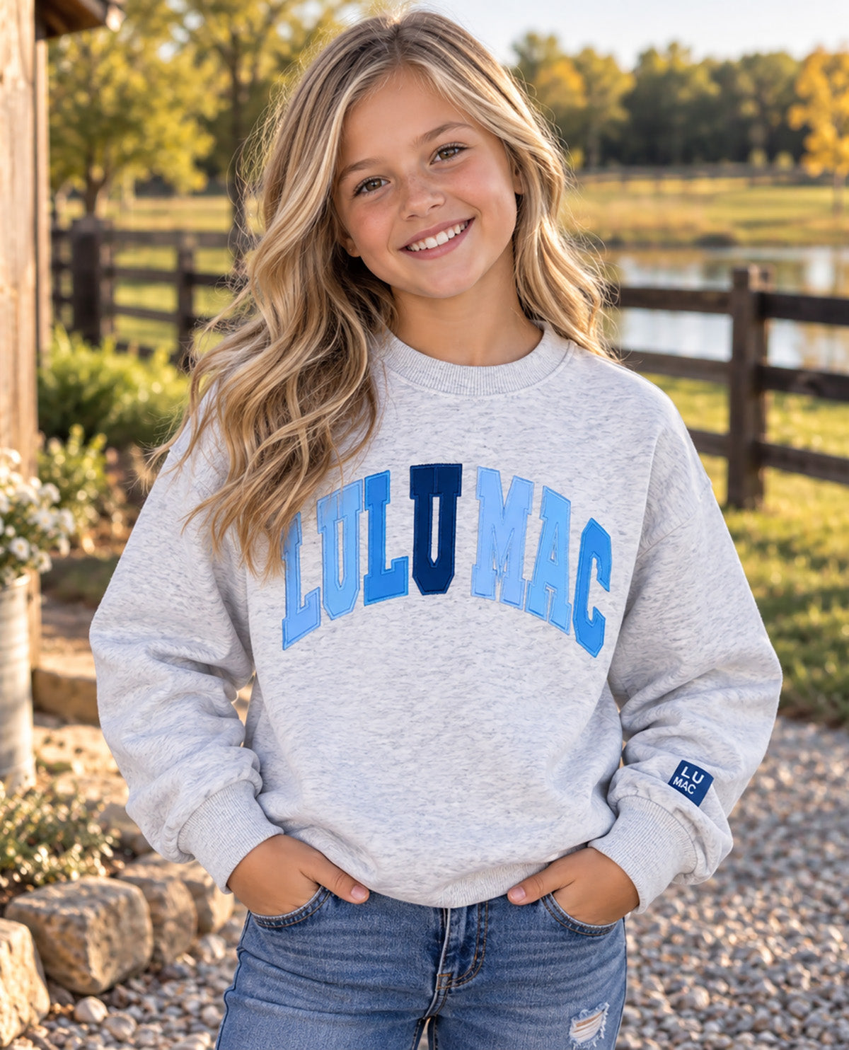A young girl smiling outdoors, wearing a light heather gray sweatshirt with 'LULU MAC' printed across the chest in blue and navy varsity-style lettering, paired with blue jeans.