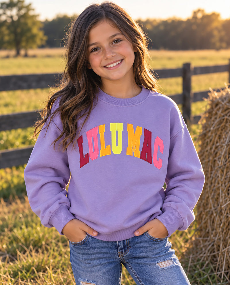A young girl smiling outdoors in a lavender crewneck sweatshirt with 'LULU MAC' printed across the chest in colorful letters, paired with blue jeans, set against a rustic farm background with a fence and hay bale.