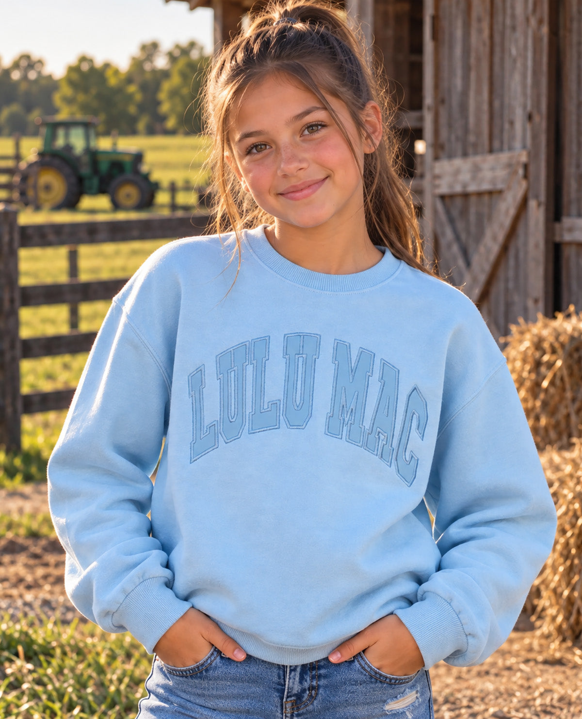 A young girl with her hair in a high ponytail stands outdoors in front of a rustic barn and fence, wearing a light blue crewneck sweatshirt with 'LULU MAC' printed in arched block letters across the chest, paired with blue jeans.
