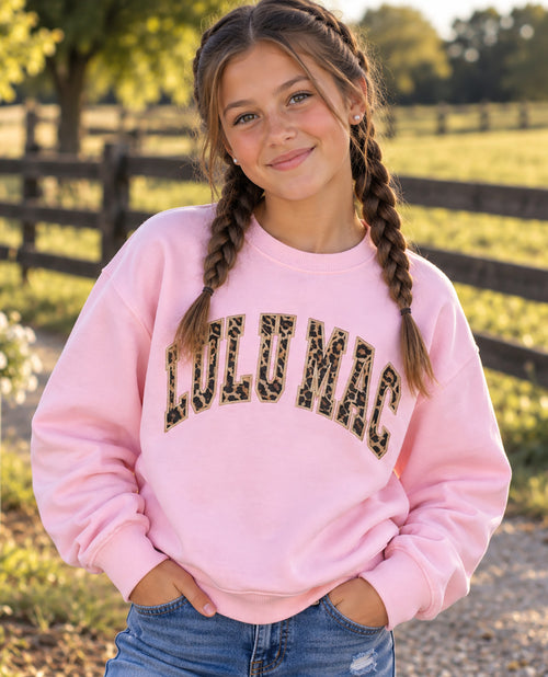 A young girl with braided hair wearing a pink crewneck sweatshirt with 'LULU MAC' printed across the chest in a leopard print pattern, standing outdoors in a sunny field.