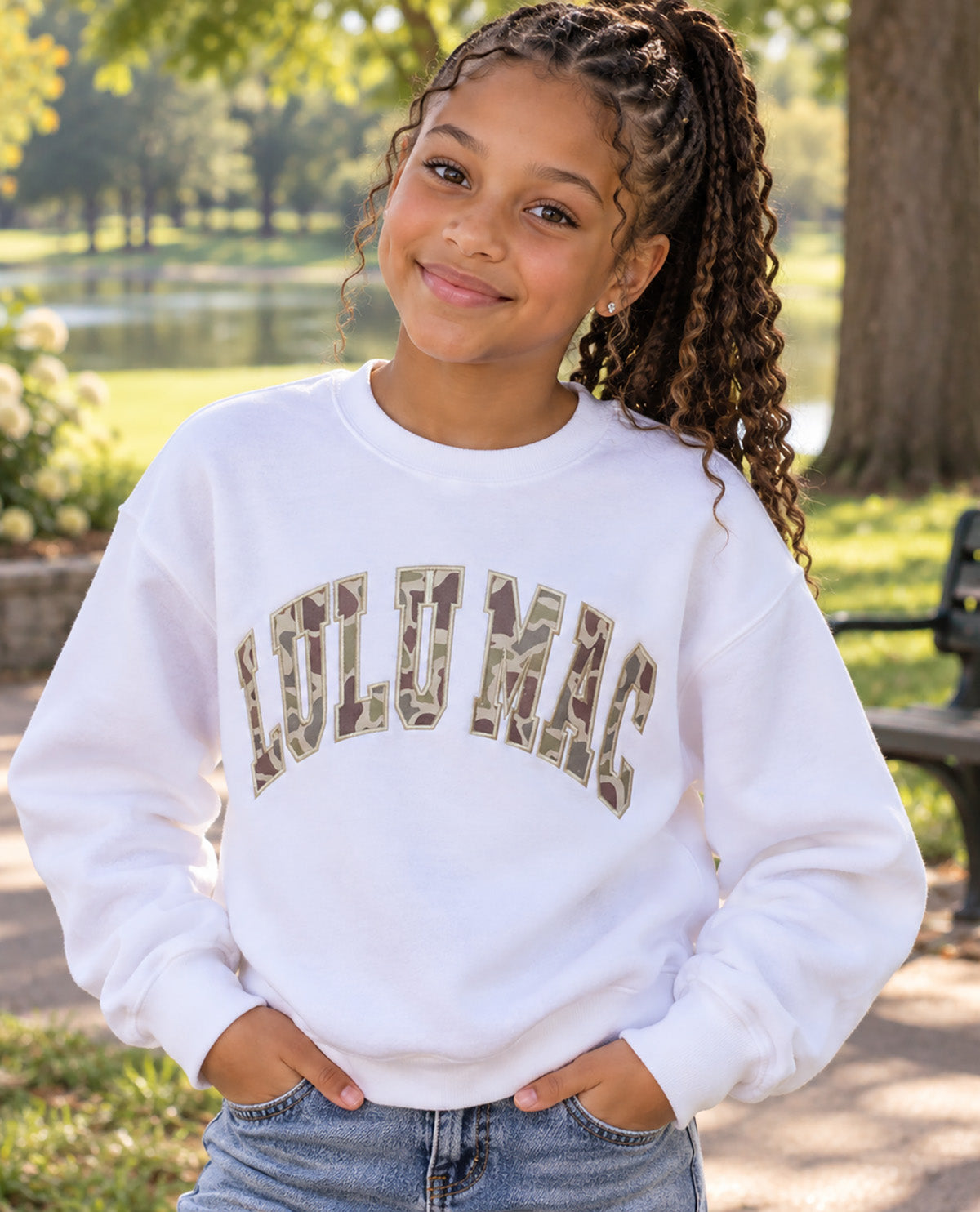 A young girl with braided hair wearing a white crewneck sweatshirt with 'LULU MAC' printed in camouflage letters, standing outdoors in a park setting.