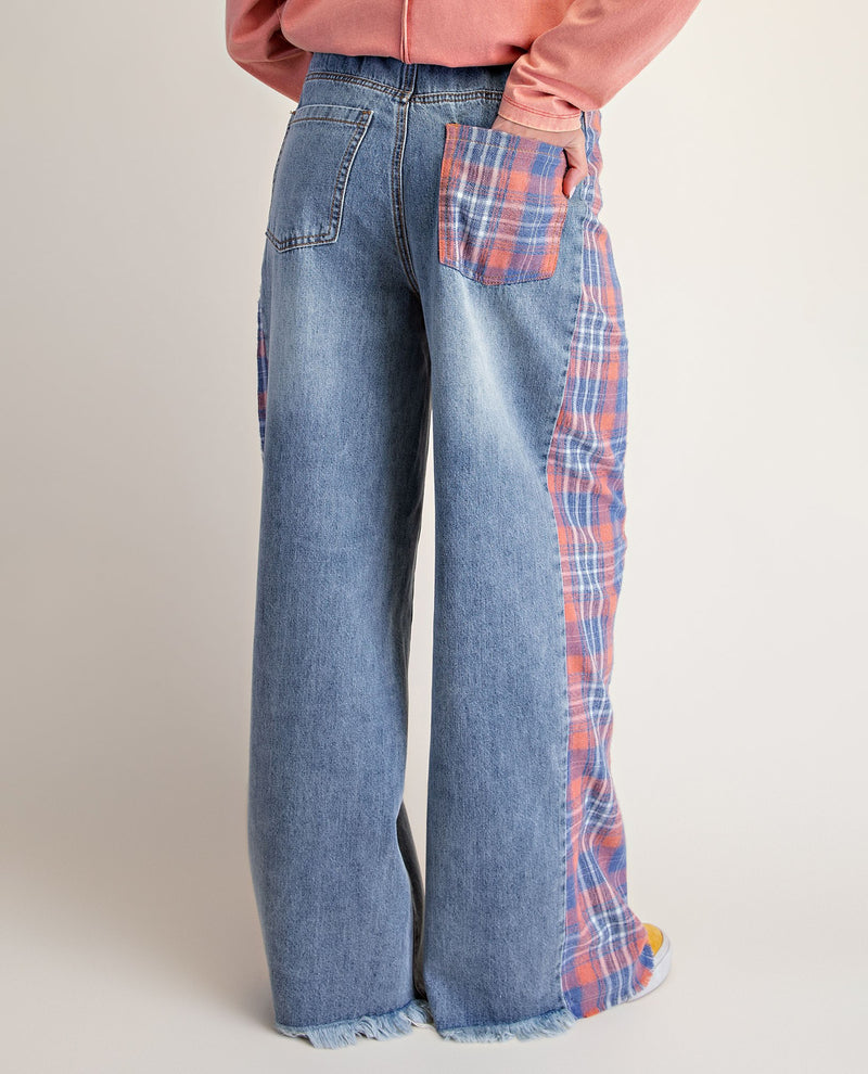 A person wearing wide-leg blue denim jeans with red and blue plaid patchwork pocket and side panel details, paired with a pink top, photographed against a plain light beige studio background.