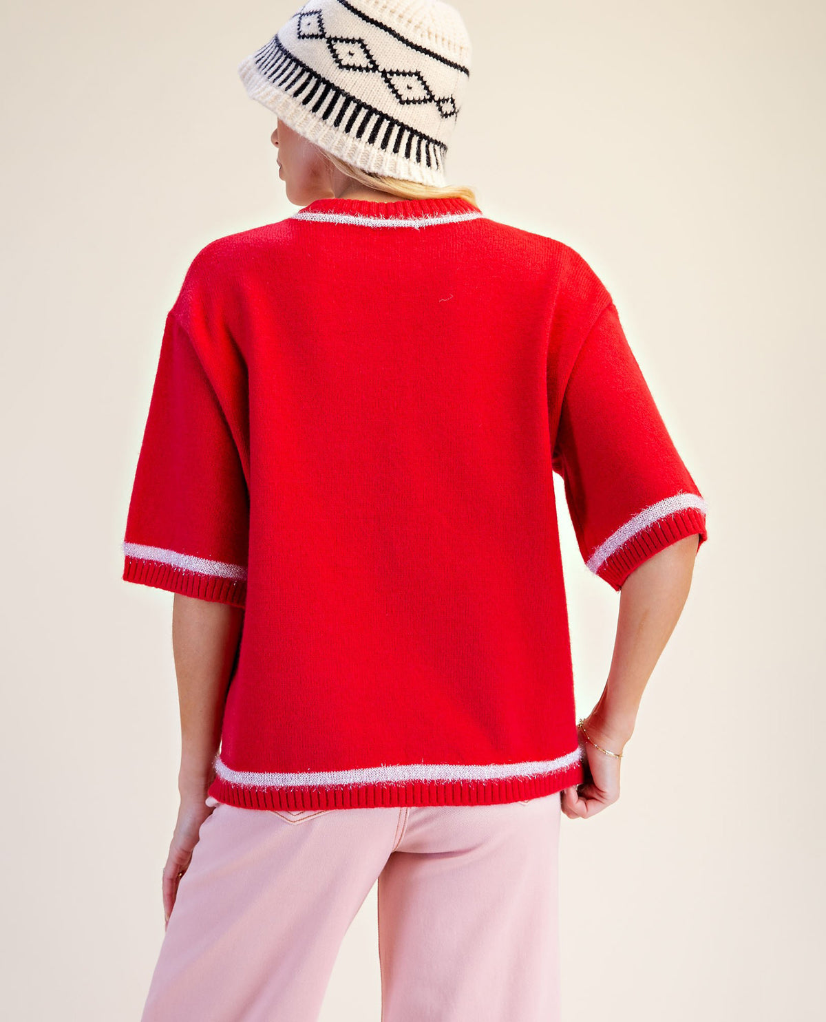 A person shown from the back wearing a bright red knit sweater with white ribbed trim at the collar, sleeve cuffs, and hem, paired with light pink pants and a cream knit beanie with black geometric patterns.