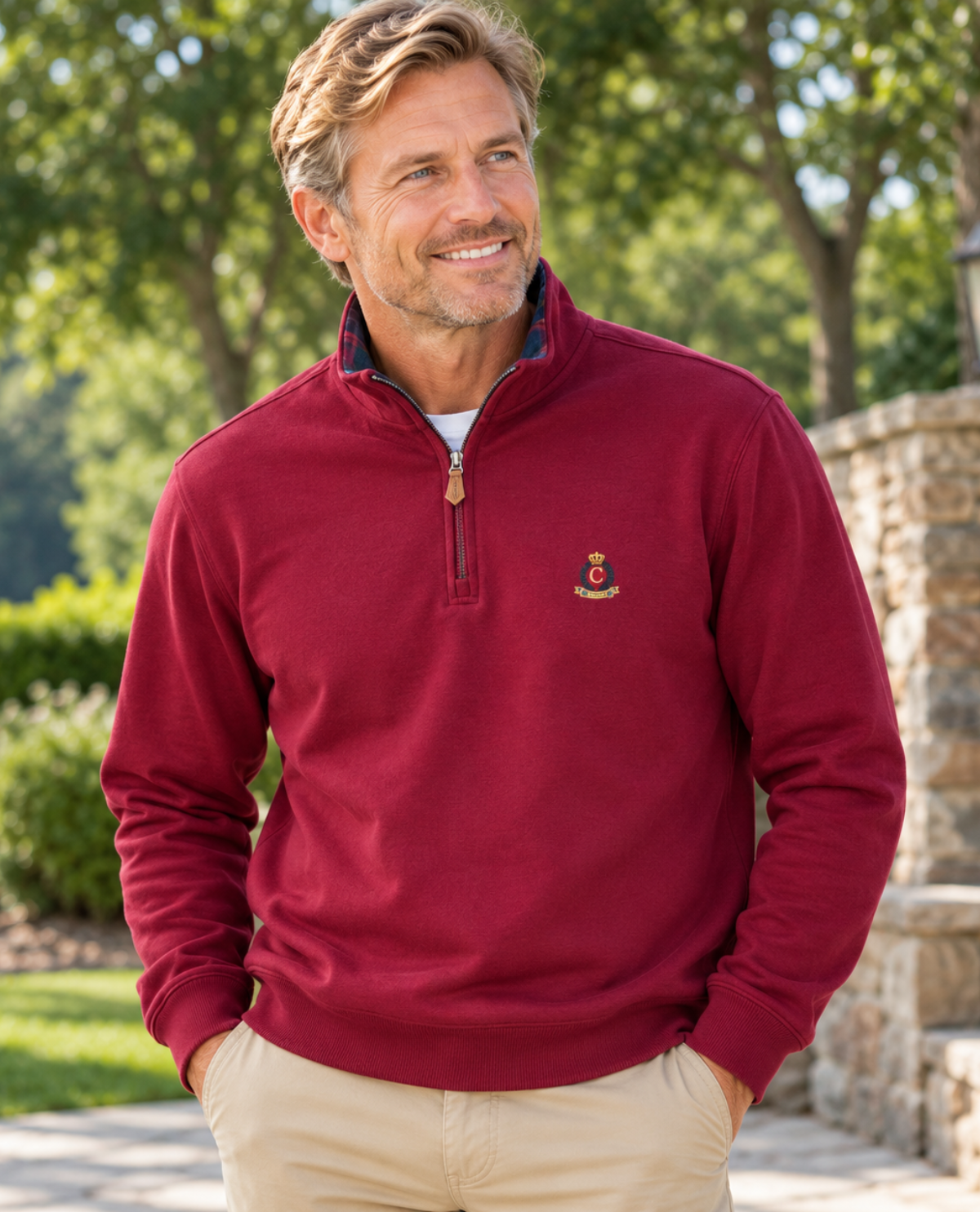 A smiling man wearing a burgundy quarter-zip pullover sweater with a small embroidered crest logo on the left chest, paired with khaki pants, standing outdoors in a garden setting.