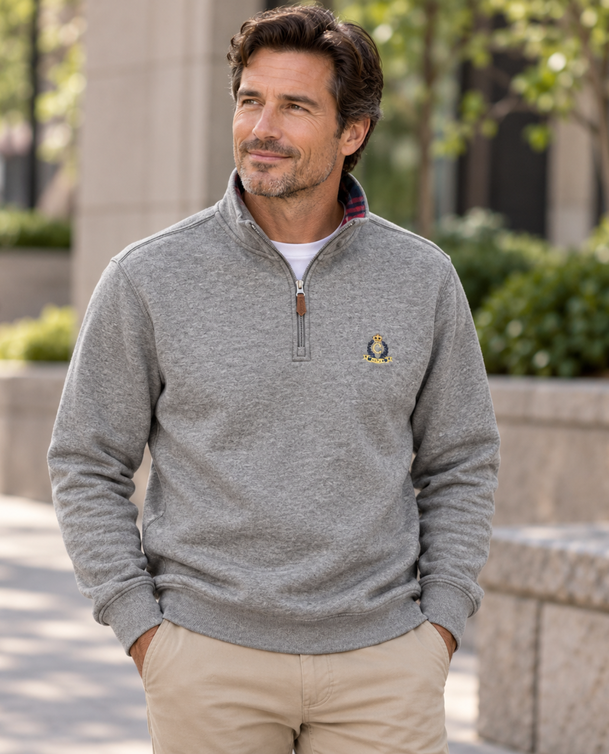 A man wearing a heather gray quarter-zip pullover sweater with a small embroidered logo on the chest, paired with khaki pants, standing outdoors in a park-like setting.
