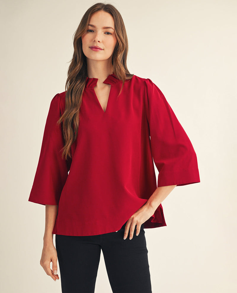 A woman modeling a vibrant red three-quarter sleeve blouse with a split V-neckline, paired with black pants, against a solid light beige background.