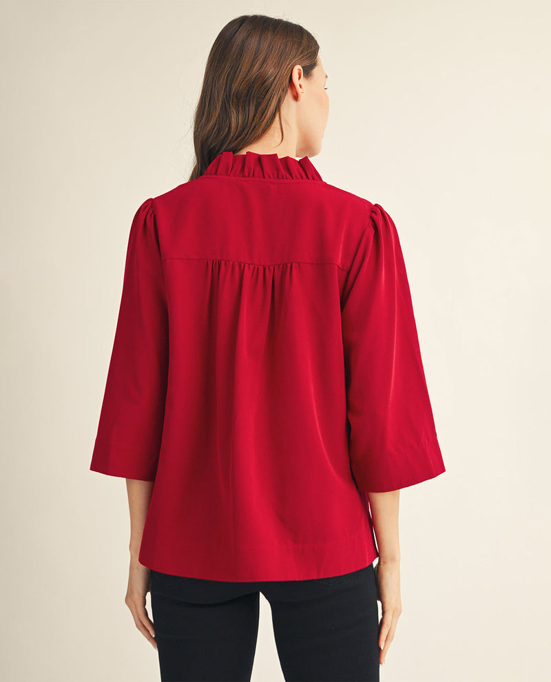 A woman viewed from the back wearing a bright red blouse with three-quarter sleeves, a ruffled high neck, and gathered detailing at the shoulders, paired with black pants against a plain light beige studio background.