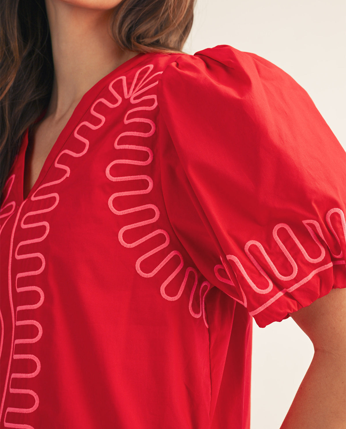 Close-up of a red blouse with puff sleeves and light pink wavy embroidered trim along the neckline and sleeve, worn by a model.
