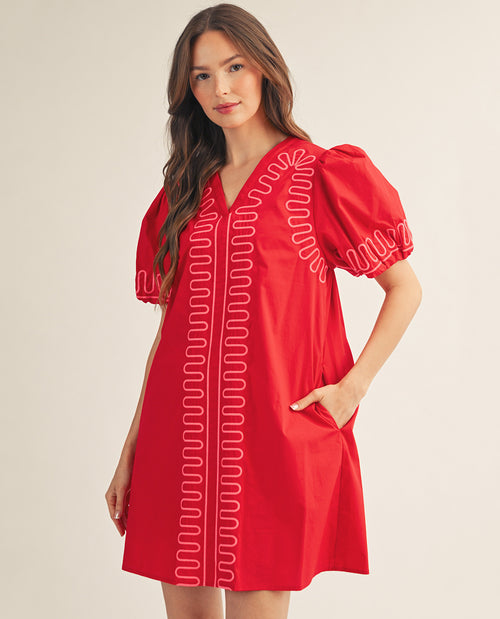 A woman modeling a bright red short-sleeve dress with white embroidered wavy line patterns down the front and on the sleeve, set against a plain beige studio background.