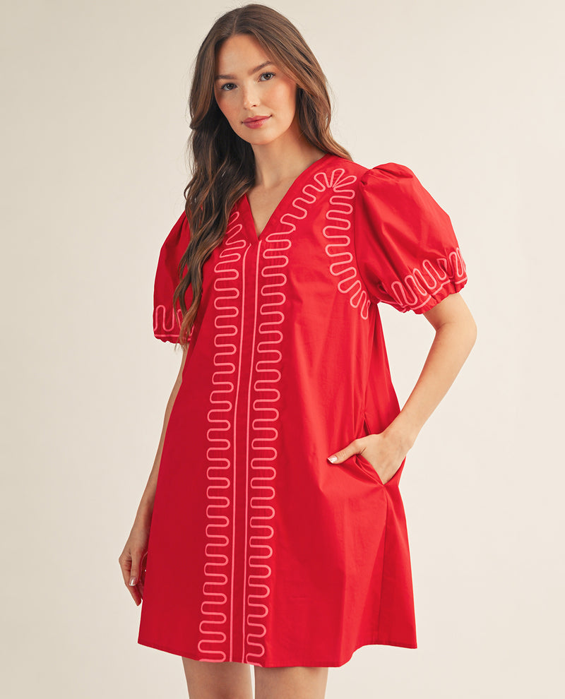 A woman modeling a bright red short-sleeve dress with white embroidered wavy line patterns down the front and on the sleeve, set against a plain beige studio background.