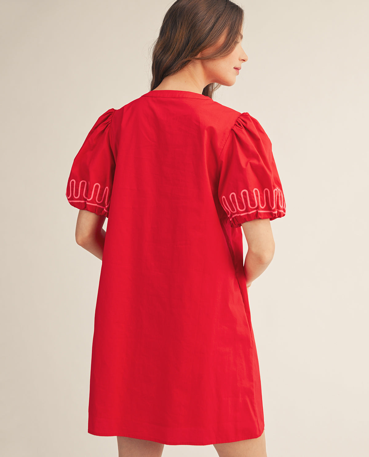 A woman wearing a bright red short-sleeve dress, shown from the back. The dress features voluminous puff sleeves with white scalloped embroidery along the cuffs, set against a plain beige studio background.
