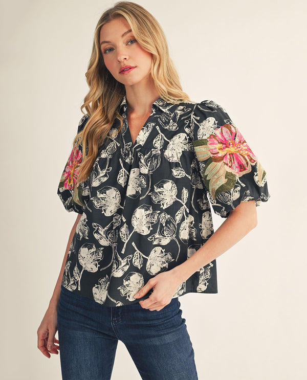 A woman modeling a short-sleeve button-up blouse with a black base and a white floral print, featuring pink and green embroidered floral details on the puff sleeves, paired with dark blue jeans against a light beige background.