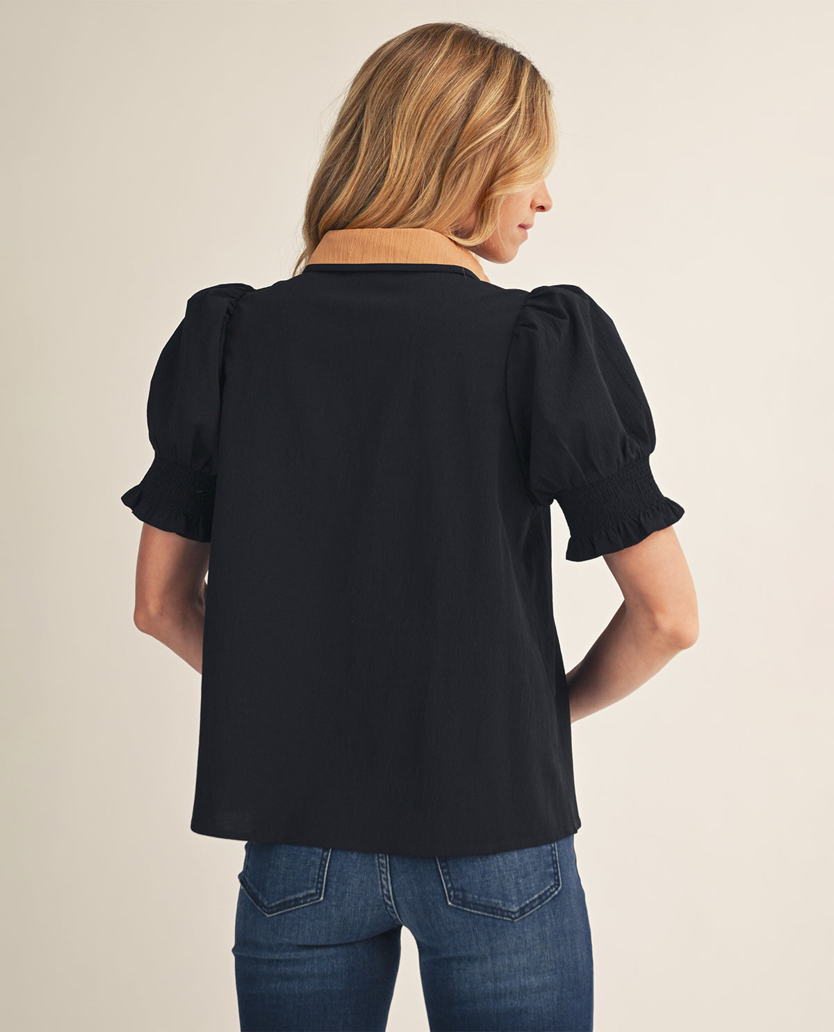 A woman shown from the back wearing a black short-sleeve blouse with puffed sleeves and a tan collar, paired with blue jeans, against a plain light beige studio background.