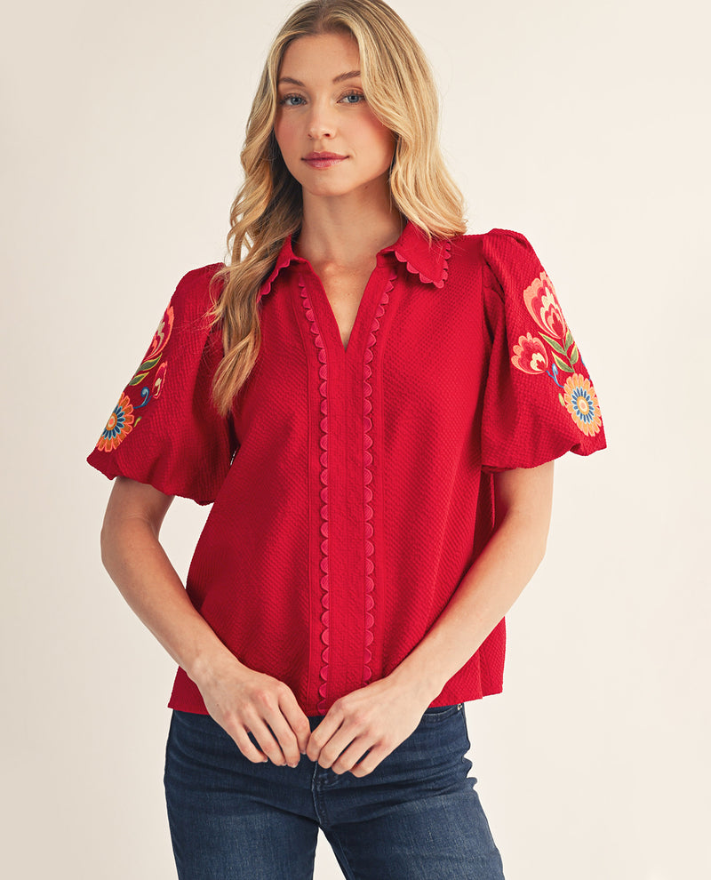 Woman wearing a red blouse with floral embroidery on sleeves against a beige background