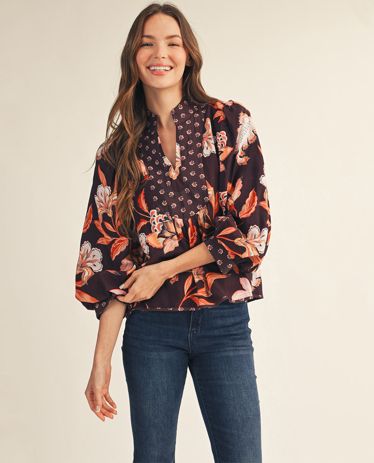A woman modeling a dark floral blouse with orange, pink, and white botanical print, paired with dark blue jeans, against a solid light beige background.