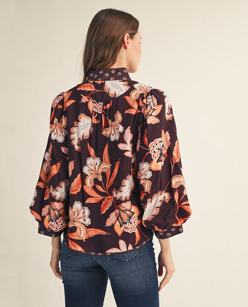 A woman shown from the back wearing a dark floral blouse with orange, pink, and cream flowers, paired with blue jeans, against a plain beige studio background.