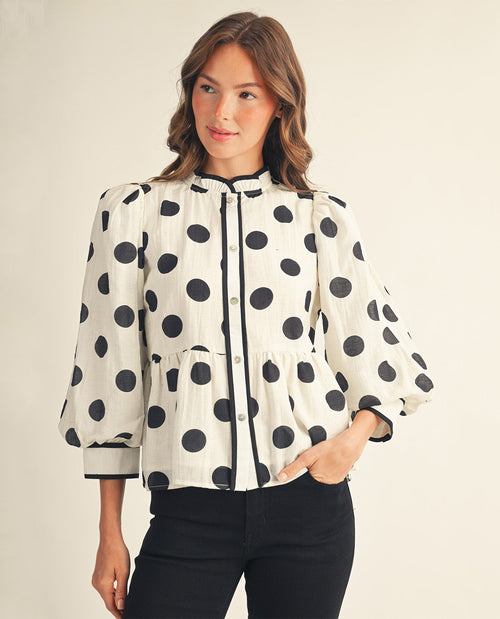 A woman wearing a white blouse with large black polka dots, black trim, and puff sleeves, paired with black pants, against a plain light beige studio background.
