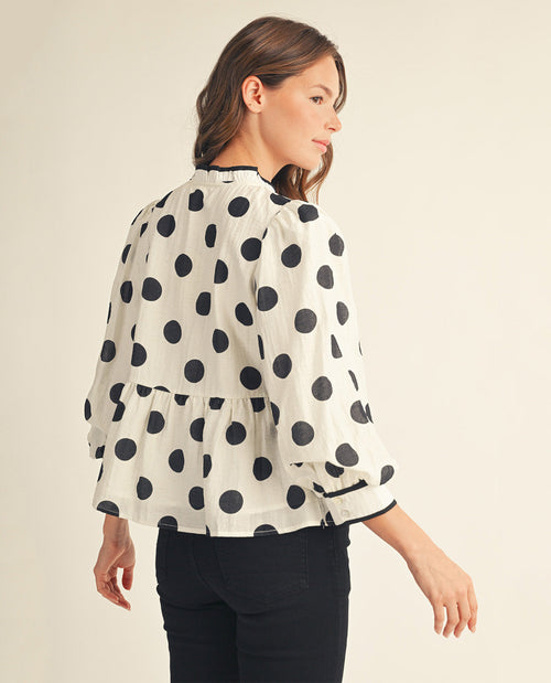 A woman shown from the back wearing a cream-colored blouse with large black polka dots, paired with black pants, against a plain light beige background.