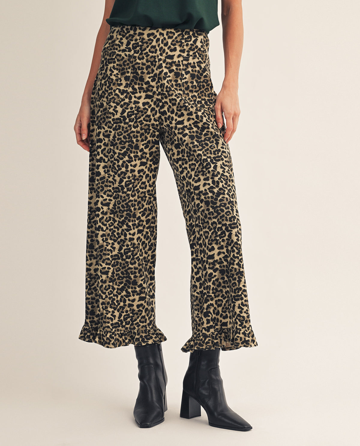 A model wearing high-waisted, wide-leg cropped pants with a beige and black leopard print, paired with black ankle boots, against a plain light beige studio background.