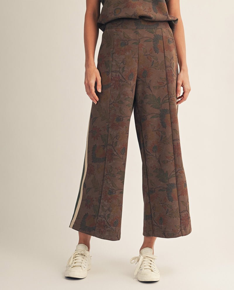 A model wearing brown wide-leg cropped trousers with a botanical jacquard pattern and a beige stripe detail along the side, paired with white sneakers against a light beige studio background.