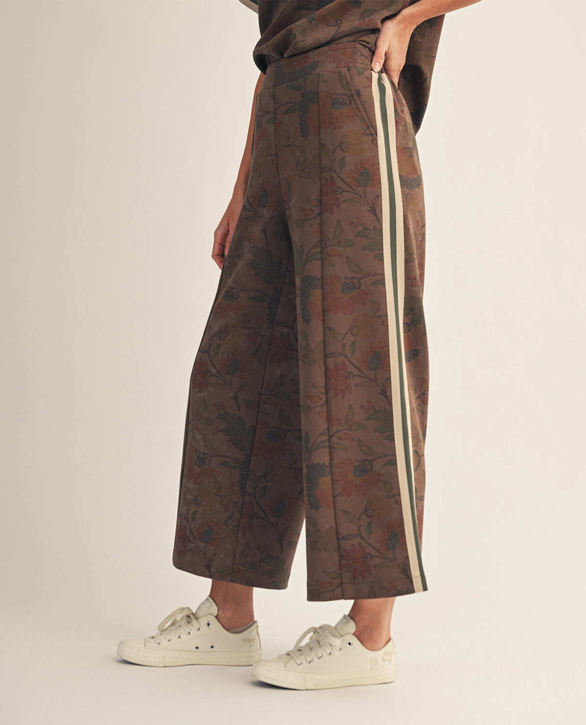 A person wearing wide-leg cropped trousers with a brown floral jacquard pattern and a cream-colored side stripe, paired with white sneakers, photographed against a plain light beige studio background.