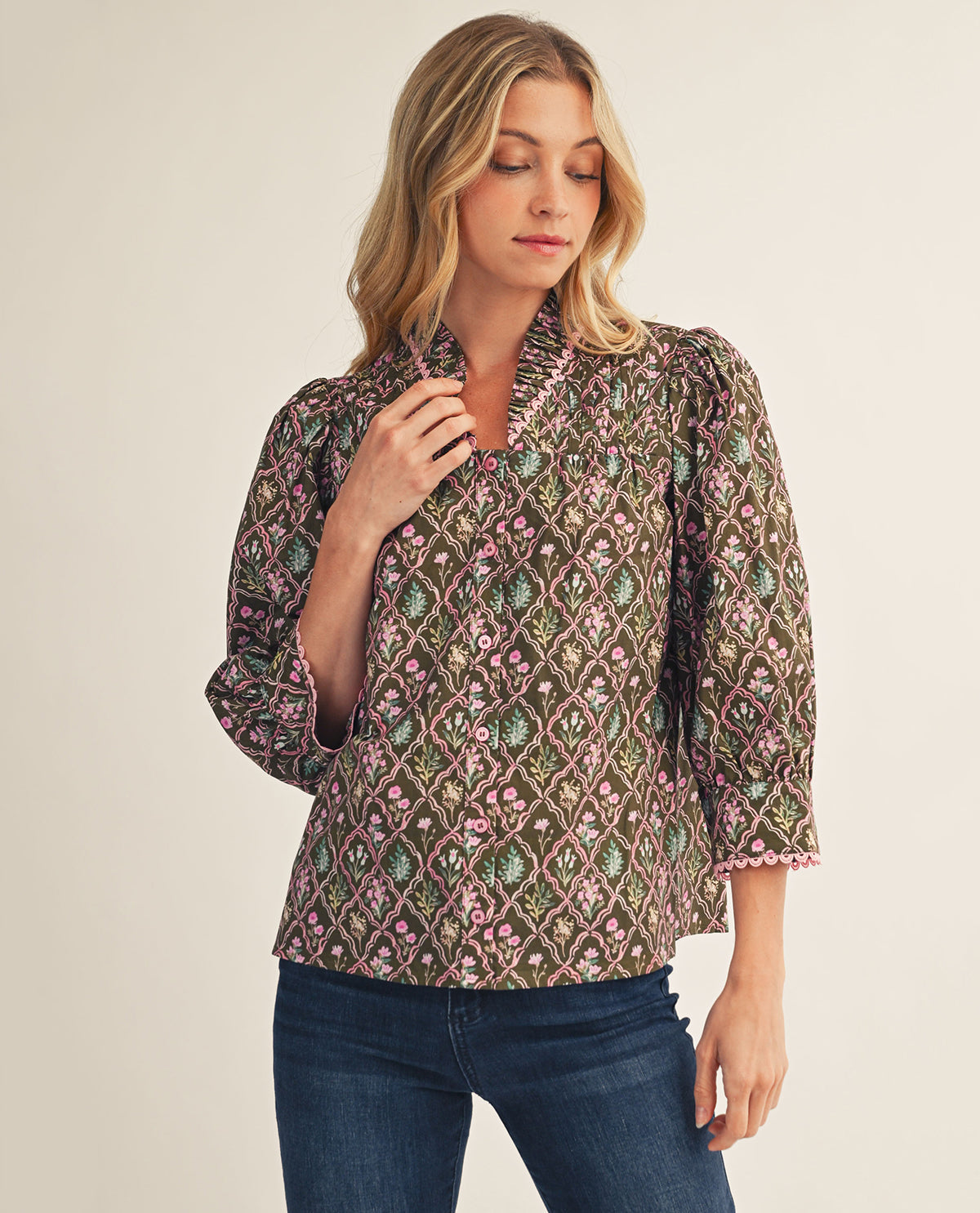 A woman wearing a patterned three-quarter sleeve blouse with dark blue jeans, standing against a plain light beige studio background.