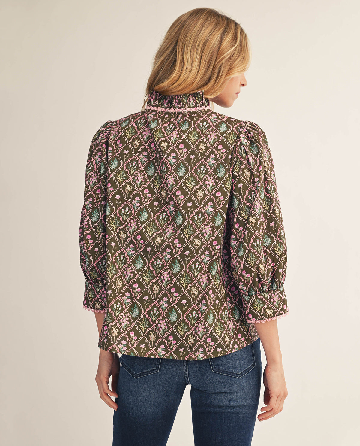 A woman shown from the back wearing a patterned blouse with three-quarter sleeves and blue jeans, photographed against a plain light beige studio background.