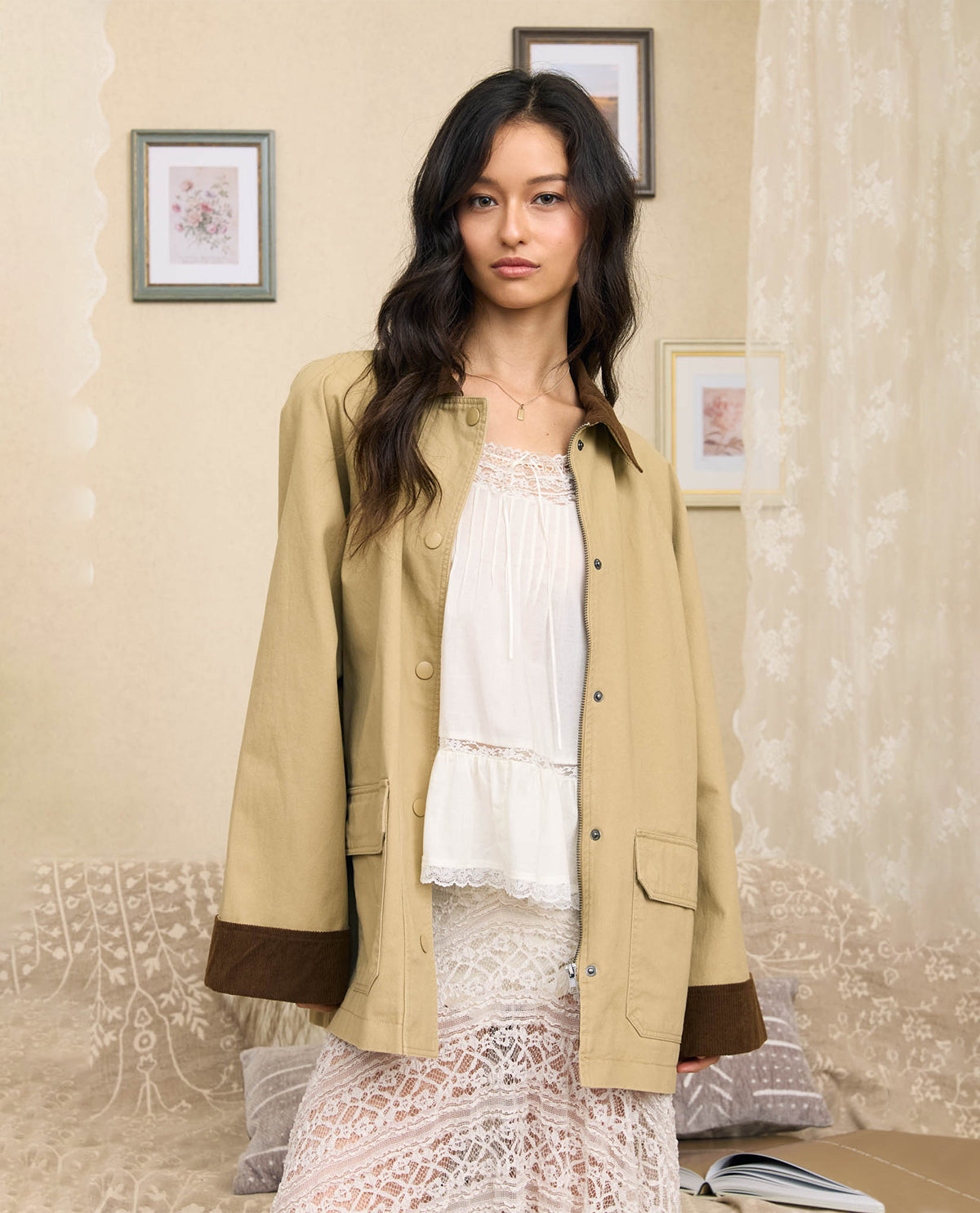 A woman stands indoors wearing a beige button-front jacket with dark brown cuff panels over a white lace top and skirt. The room has cream walls, framed art, sheer curtains, and a patterned sofa.
