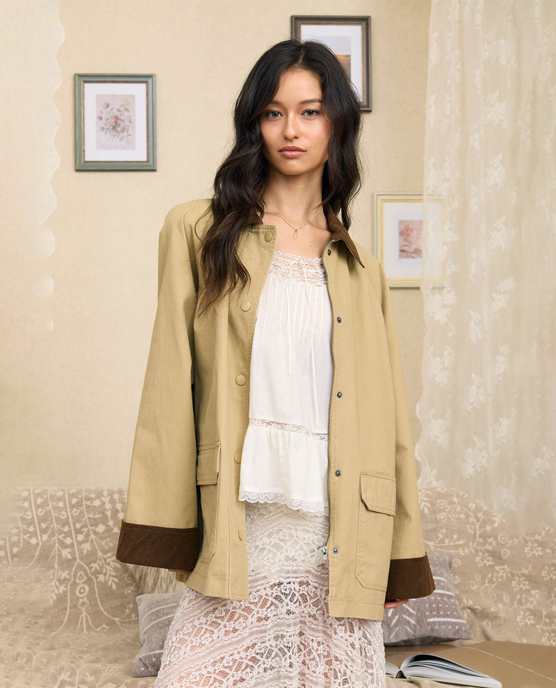 A woman stands indoors wearing a beige button-front jacket with dark brown cuff panels over a white lace top and skirt. The room has cream walls, framed art, sheer curtains, and a patterned sofa.
