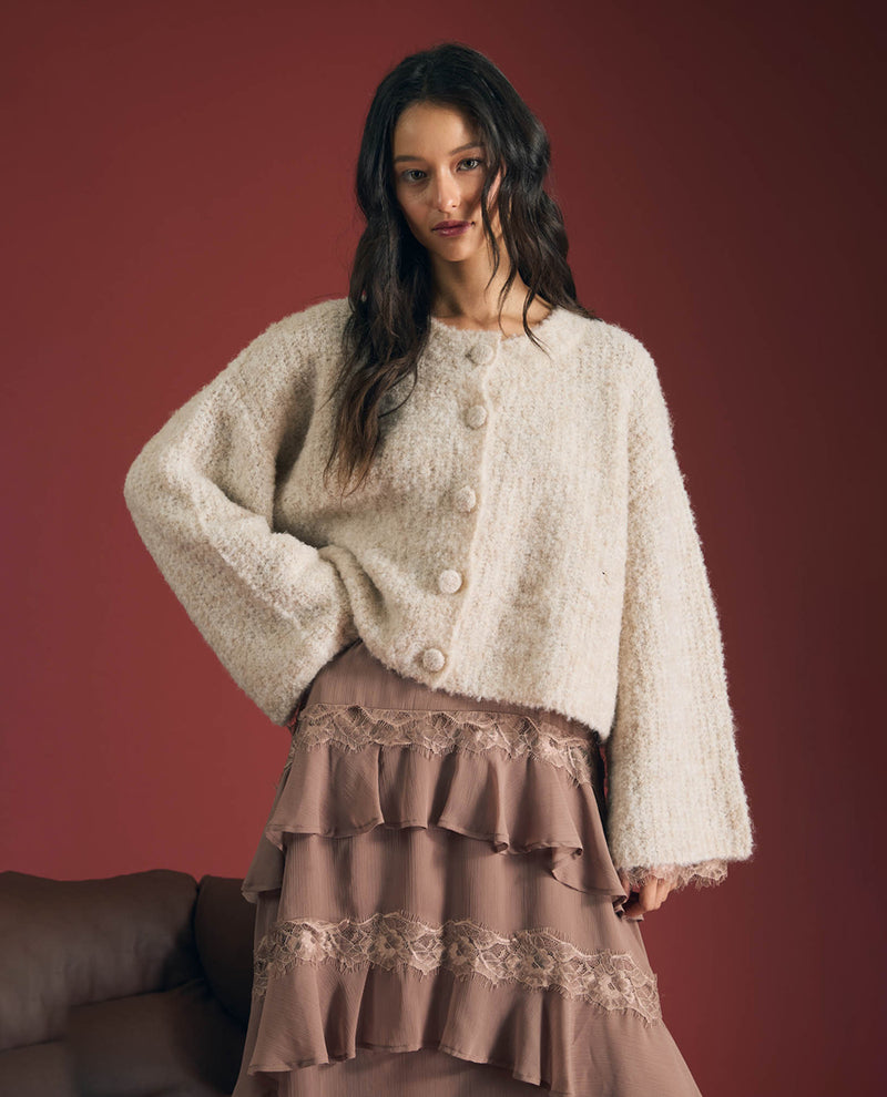 A woman models a fuzzy cream-colored cropped cardigan with large buttons, paired with a brown ruffled tiered skirt, set against a solid dark reddish-brown background.