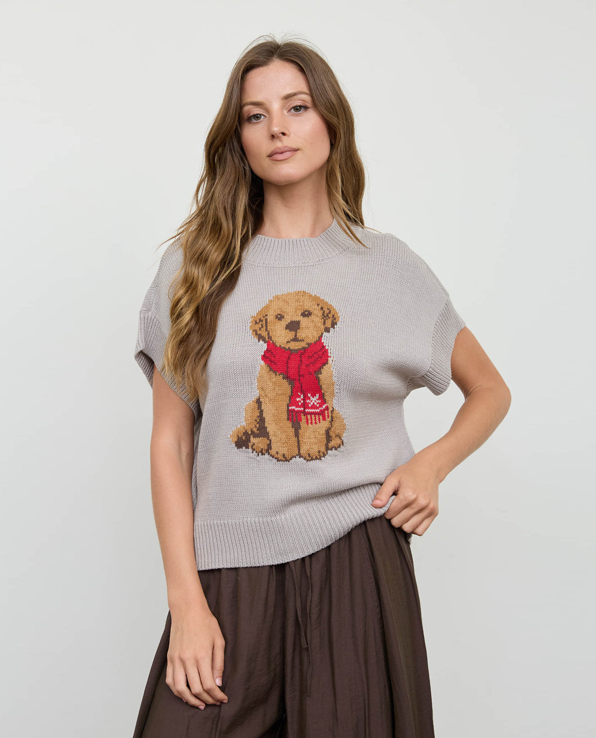 A woman wearing a light gray knit sweater with a knitted graphic of a golden retriever puppy wearing a red scarf, paired with dark brown wide-leg pants, against a light gray studio background.