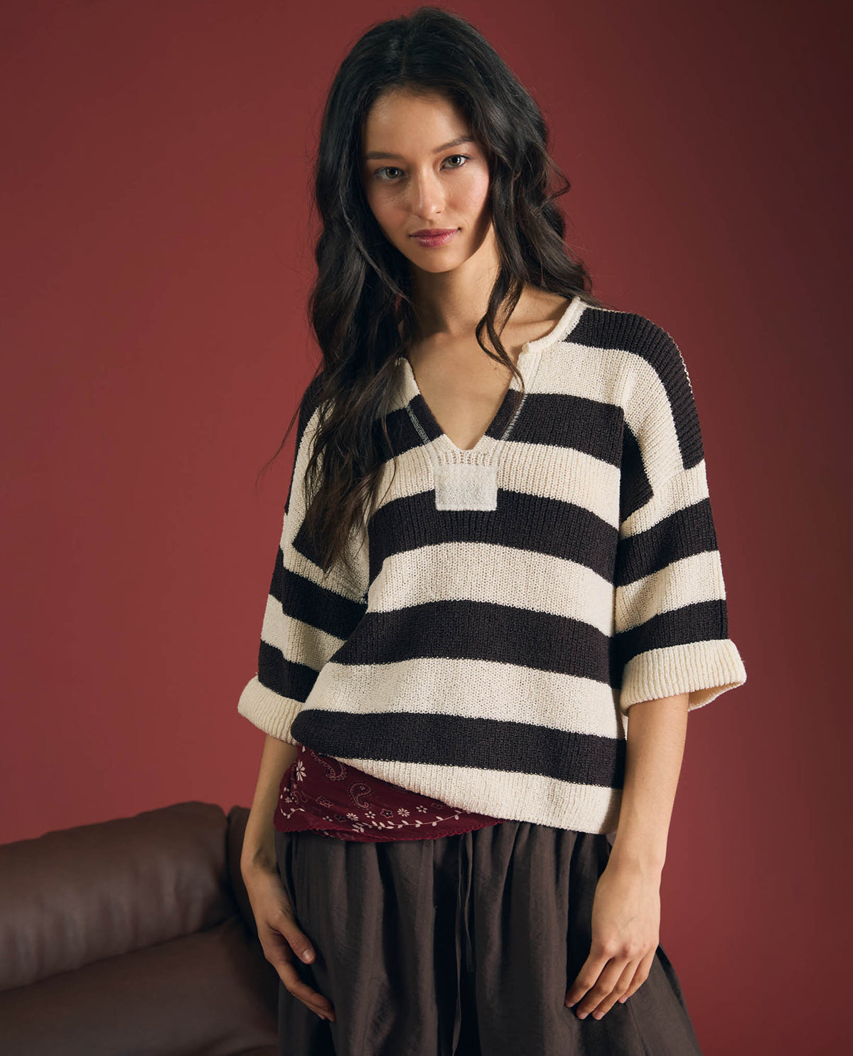A woman wearing a black and cream striped knit sweater with elbow-length sleeves and a V-neck, paired with dark brown pants and a burgundy lace-trimmed skirt, posed against a solid dark red-brown studio background.