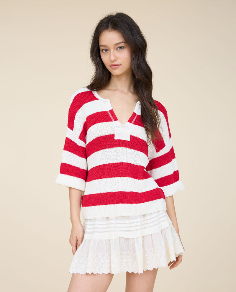 A woman modeling a red and white horizontally striped knit sweater with three-quarter sleeves, layered over a white pleated mini skirt, against a solid beige background.