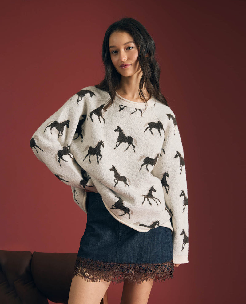 A woman modeling a cream-colored knit sweater with a black horse pattern, paired with a dark denim skirt, against a solid dark red background.