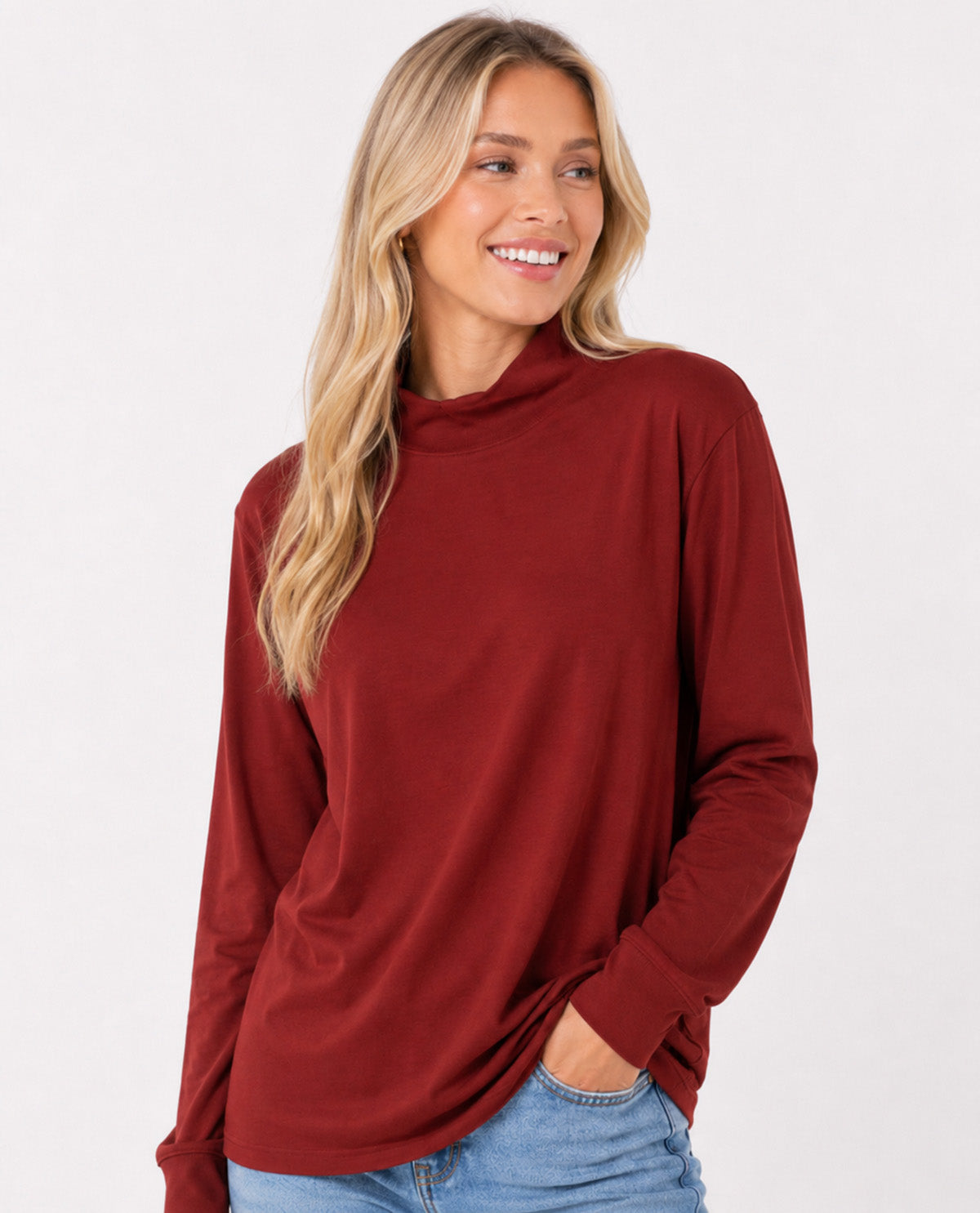 A woman with blonde hair wearing a long-sleeve, high-neck turtleneck top in a deep burgundy red, paired with light blue jeans, against a plain white studio background.