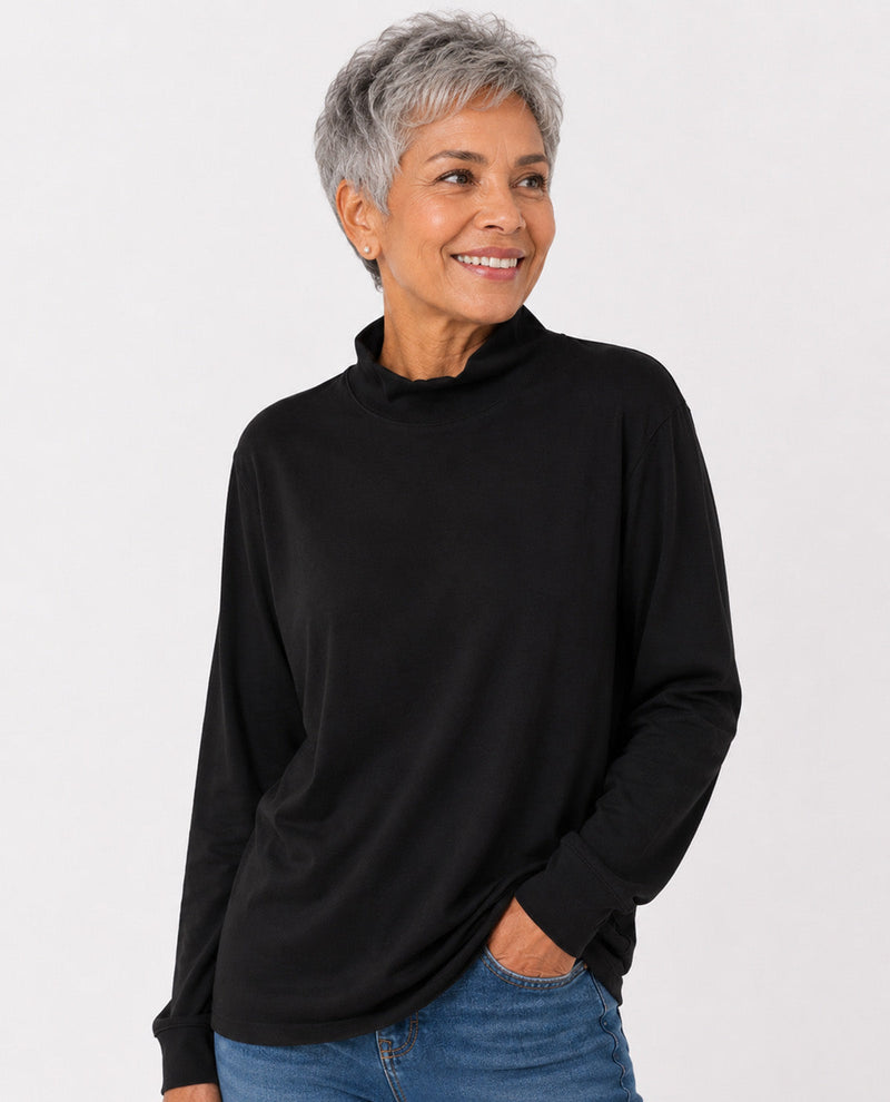 A woman with short gray hair wearing a black long-sleeve mock neck top and blue jeans, standing against a light gray studio background.