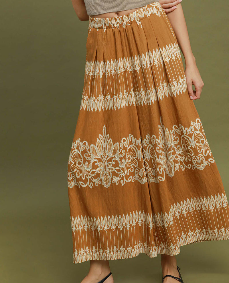 A wide-leg pleated skirt in a warm rust-orange color with intricate cream-colored floral embroidery and geometric patterns, shown on a model against a solid olive green background.