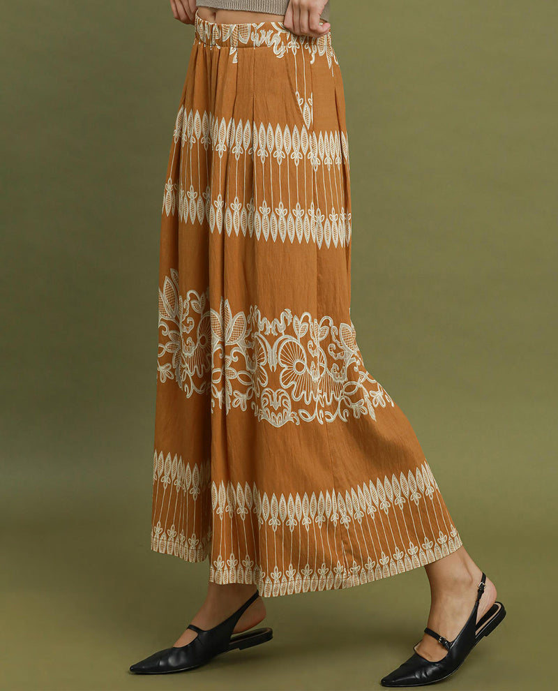 A person wearing a wide-leg pleated maxi skirt in a warm camel brown with cream-colored embroidered patterns, paired with black flat Mary Jane shoes, set against a solid olive green background.