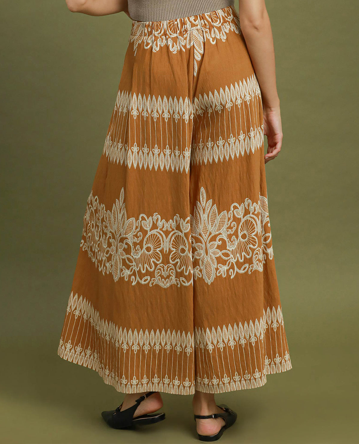 A person wearing a wide-leg, rust-orange maxi skirt with intricate cream-colored floral embroidery and geometric borders, paired with black flat sandals against a solid olive green background.
