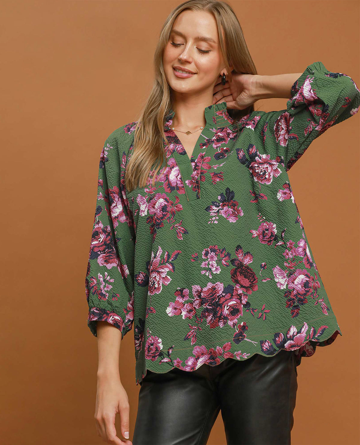 A woman wearing a green long-sleeve blouse with a pink and purple floral pattern, paired with black leather pants, posed against a solid brown studio background.
